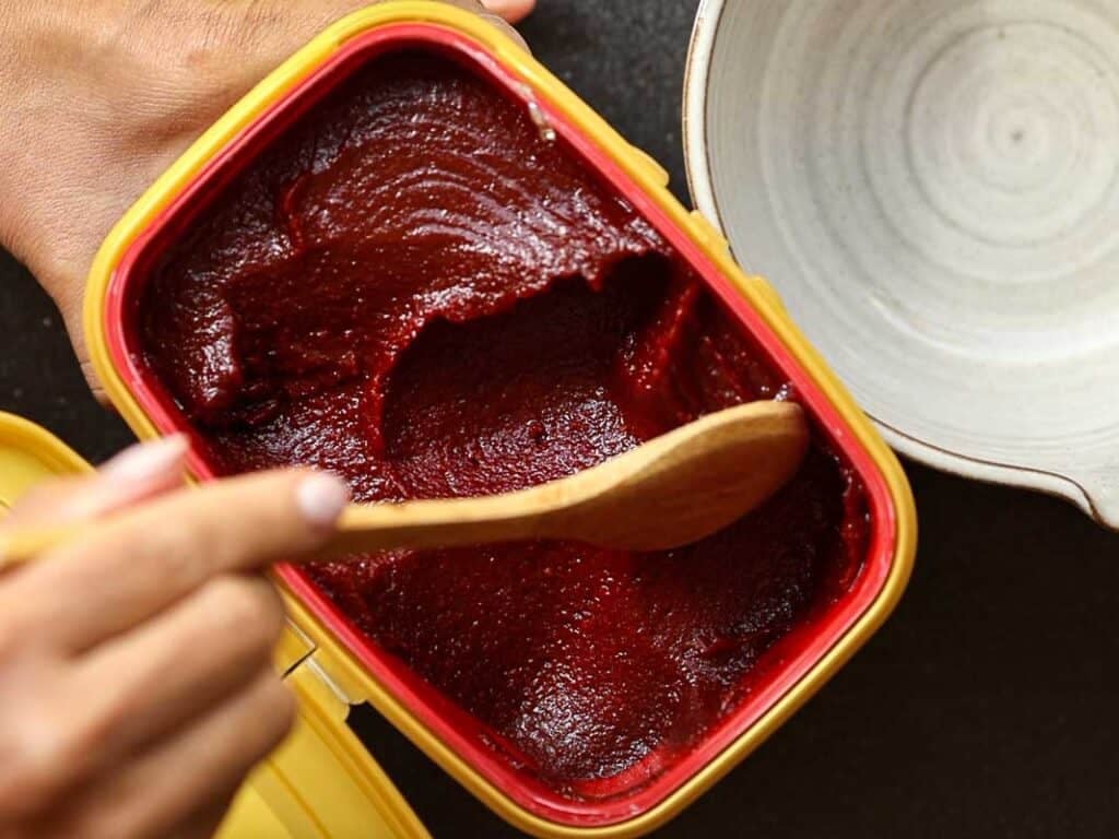 A hand holds a wooden spoon scooping dark red paste from a rectangular yellow container. A light-colored bowl is partially visible beside the container. The paste has a smooth texture.
